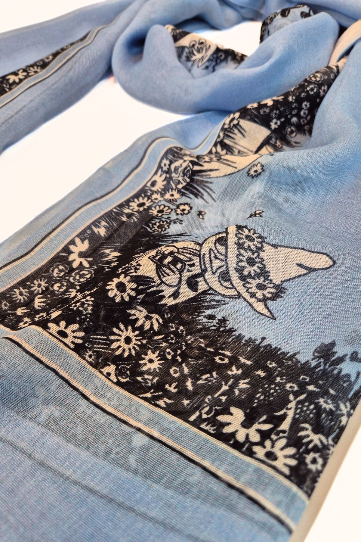 Lähikuvassa sinisen Muumi-huivin kesäinen kuvio ja Nuuskamuikkunen, ja hengittävä lyocell-materiaali